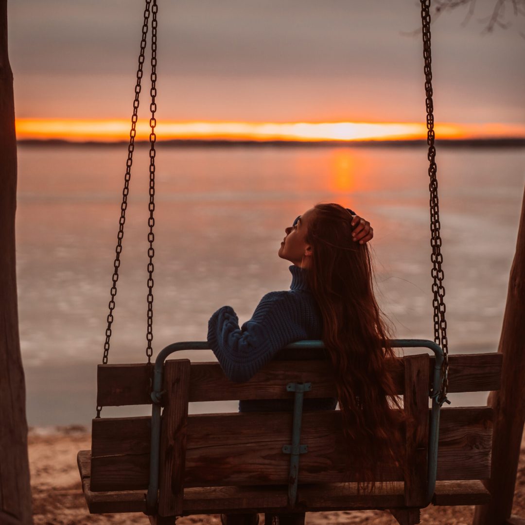 Woman enjoying time relaxing by the beautiful lake at sunrise. Girl on a wooden swing